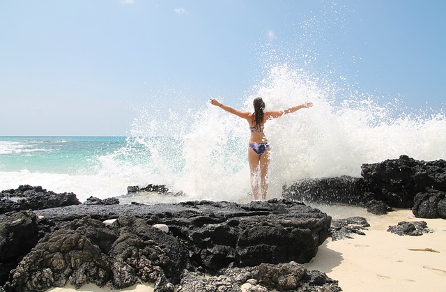 A woman standing on a rocky beach embracing the waves in her bikini